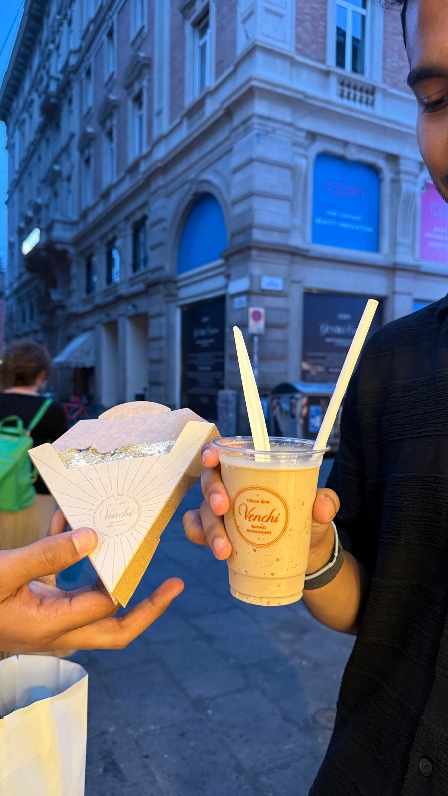 A gelato cone and an iced drink from Venchi held up on a street in Bologna at dusk
