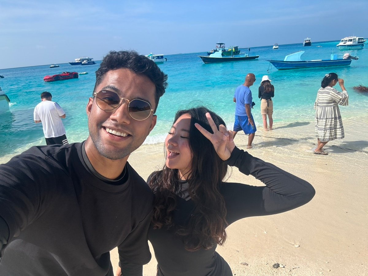 Alyssa and a friend taking a selfie on a beach with bright turquoise water and boats behind them